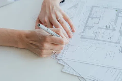 Person's hands sketching architectural floor plans on paper at a white desk with a pencil and phone nearby