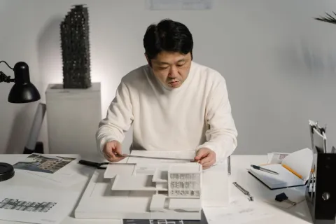Architect studying architectural model and blueprints at desk with design tools and documents in office.