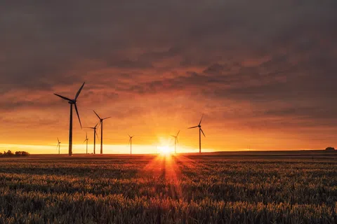 Wind turbines in a field at sunset with dramatic orange sky and sun rays over the horizon.