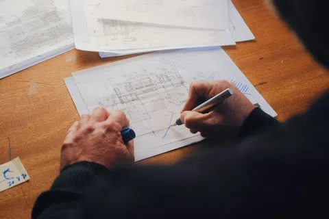 Person working on architectural blueprints with pen at wooden desk near markers container.
