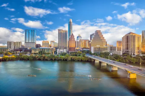 Austin city skyline with high-rise buildings, bridge over the river, and boats rowing on the water under blue sky.