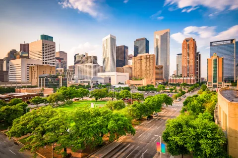 Daytime city skyline with tall buildings, green trees, and clear blue sky with clouds over a busy urban area.