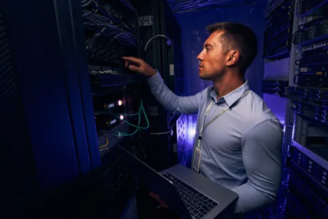 IT technician in a server room working on network equipment with a laptop under blue lighting.