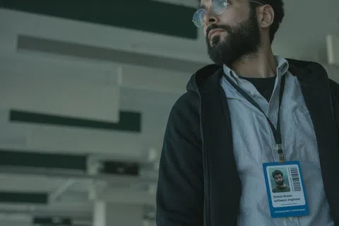 Bearded man wearing glasses and ID badge in an office setting looking to the side.