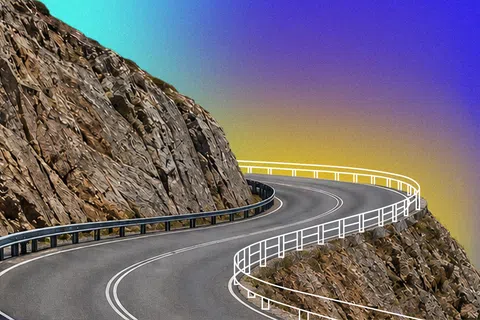 Winding mountain road with guardrails curving around rocky cliffs under a gradient sky.