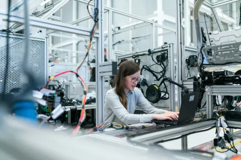 Engineer working on a laptop in a modern automotive testing lab with car parts and wiring harnesses.