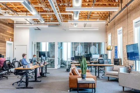 Modern open-plan office with exposed ceiling, wooden walls, desks, computers, and people working.