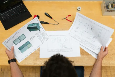 Designer reviewing technical blueprints and 3D models at a wooden desk with laptop and tools.