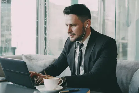 Young businessman in a suit working on laptop with earphones and coffee in a modern cafe by the window.