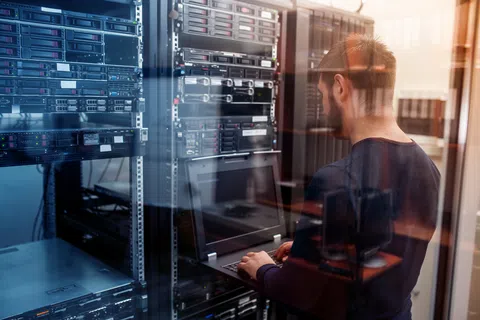 IT professional working on a laptop inside a server room with racks of network equipment and servers.