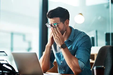 Tired man in glasses rubbing eyes while working on laptop in modern office environment.