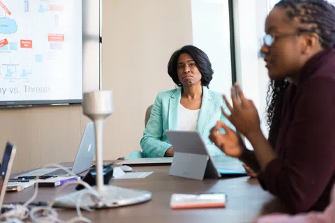 Two women engaged in a business meeting with laptops and a digital presentation on strategy versus threats visible.