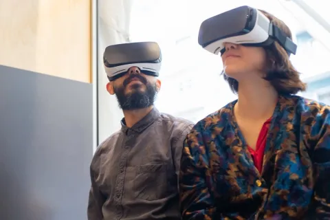 Two people wearing virtual reality headsets sitting indoors near a window experiencing VR technology.