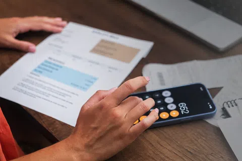 Person calculating bills using phone calculator and holding an invoice on a wooden desk with laptop and receipts