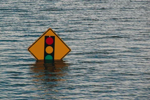 Flooded traffic signal warning sign partially submerged in murky floodwater under overcast sky.
