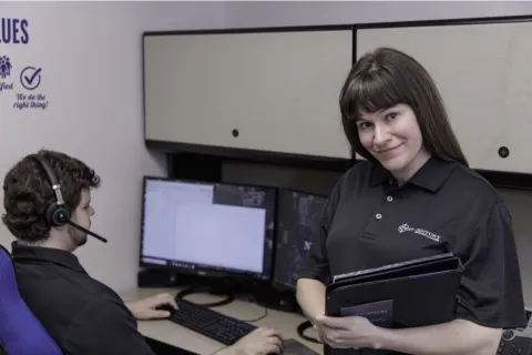 Woman holding a notepad smiling. Man behind her sitting at a desk with two monitors and a headset. Both are employees of Next Century Tech