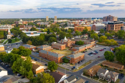 Aerial view of a cityscape with residential homes, brick buildings, green trees, and a partly cloudy sky at sunset.