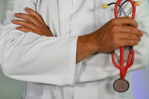 Doctor in white coat holding a red stethoscope with arms crossed against a plain background