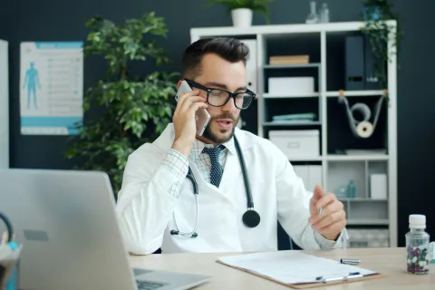 Male doctor in white coat wearing glasses talks on phone at desk with laptop and medical supplies.