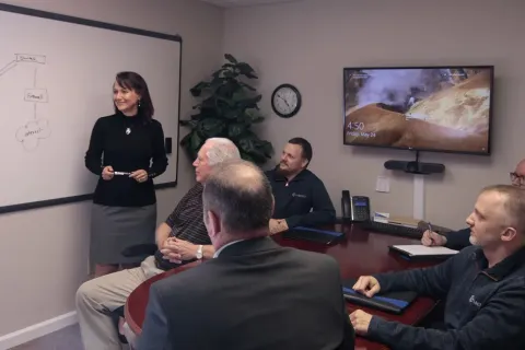 A woman standing at a whiteboard in a conference room. Five men are sitting around the table.