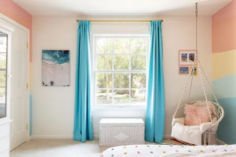 Bright bedroom with large window, blue curtains, hanging chair with pink pillow, and pastel wall stripes