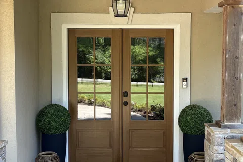 Wooden double front doors with glass panels framed by beige walls, brick flooring, and decorative plants.