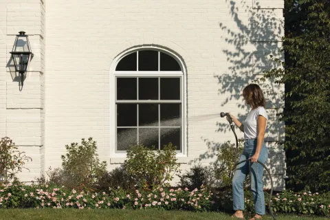 Woman watering a flower garden with a hose in front of a white brick house with an arched window.