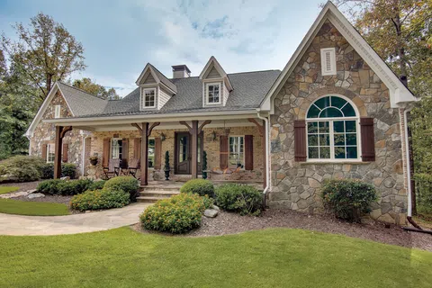 Stone and brick house with a covered porch, dormer windows, arched window, and landscaped front yard under a cloudy sky