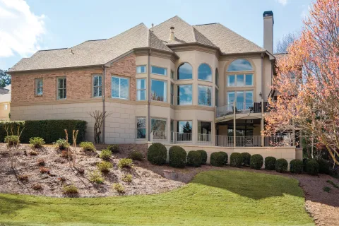 Large two-story house with beige brick exterior, multiple large windows, manicured lawn, and sparse landscaping under blue sky