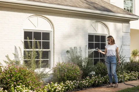 Woman watering garden flowers with hose in front of white brick house on sunny day