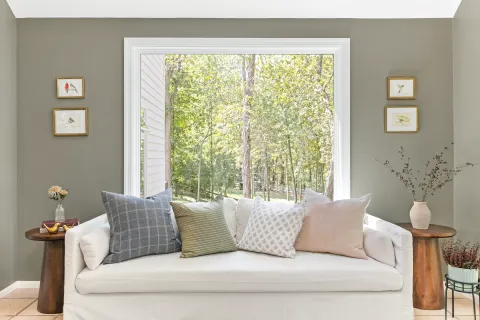Cozy white sofa with patterned pillows by large window overlooking green forest in bright living room.
