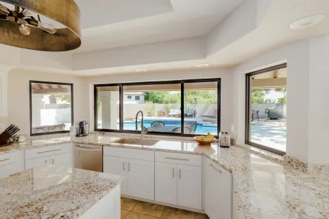 Bright kitchen with granite countertops and white cabinets overlooking a backyard pool through large windows