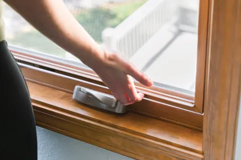 Hand opening a wooden window with a silver lock and mesh screen on a sunny day.