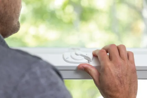 Person locking or unlocking a white window latch with blurred green outdoor background