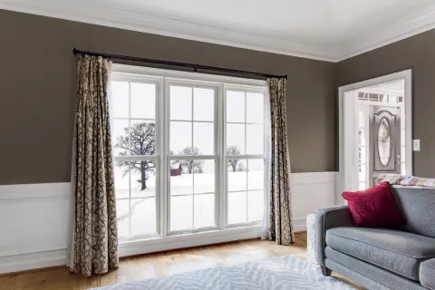 Living room with large window showing snowy landscape, gray sofa, patterned curtains, and hardwood floor.