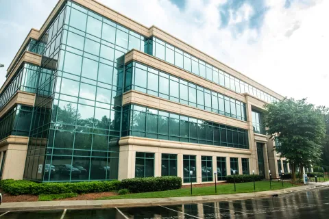Modern office building with large glass windows and beige stone accents reflected on a wet street on a cloudy day