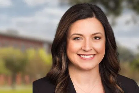 Professional woman smiling with a modern office building, trees, and water fountains in the background under a cloudy sky.