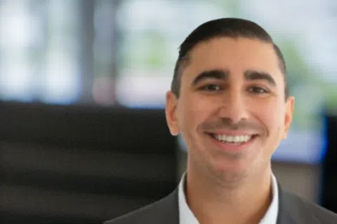 Smiling man in business suit with blurred office conference room and large windows in background.