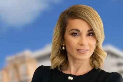 Confident businesswoman stands with folded arms in front of blurred city buildings under a blue sky.