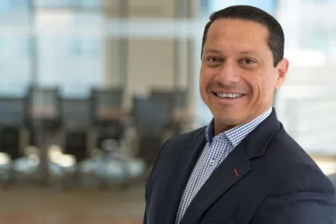 Smiling businessman in navy suit and checkered shirt in a modern office with large windows and chairs.
