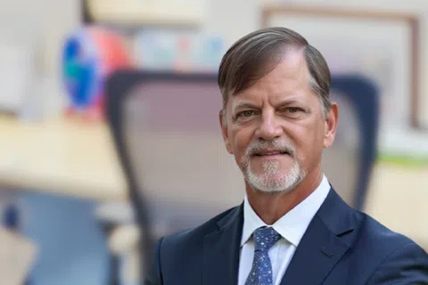 Confident businessman in suit standing with arms crossed in a modern office with empty workstations.