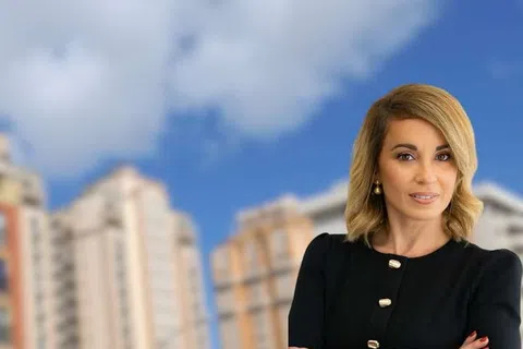 Confident businesswoman stands with folded arms in front of blurred city buildings under a blue sky.