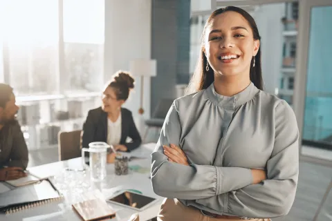 Confident young woman standing with crossed arms in modern office with colleagues talking in background
