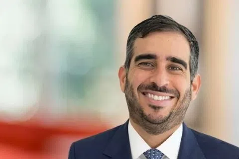 Smiling professional man in suit standing in a bright office lobby with orange seating and large windows.