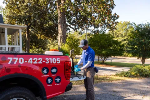 Pest control worker preparing equipment at the back of a red service truck in a residential neighborhood.