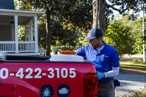 Pest control technician in blue uniform working at rear of red service truck with phone number on side.