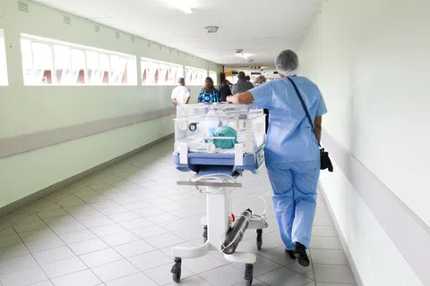 Nurse in blue scrubs pushing a neonatal incubator down a hospital corridor with medical staff ahead