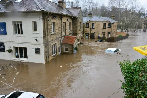 Floodwaters surround stone houses and partially submerge cars on a residential street during heavy flooding.