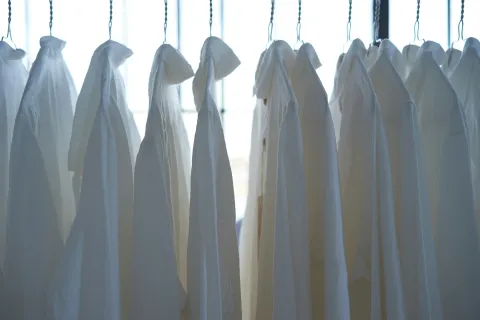 White dress shirts hanging in a row on hangers in front of a bright window.