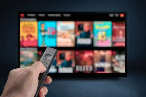 Hand holding remote control pointed at TV screen displaying blurred streaming service interface with various shows.